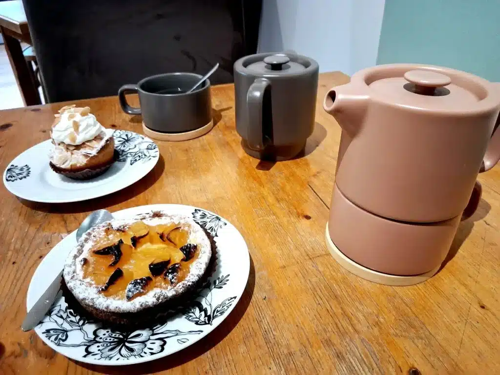 Table en bois avec une théière, des tasses et des pâtisseries artisanales, incluant une tarte aux fruits caramélisés et un gâteau aux amandes avec chantilly. ​​