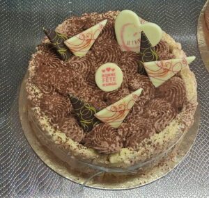 Entremets rond chocolaté, parsemé de volutes de crème cacao, décoré de cônes et plaques en chocolat blanc “Bonne Fête Maman”