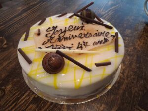 Gâteau rond glacé blanc, nappé de filet de coulis jaune, orné de bâtonnets de chocolat et d’une plaque en sucre “Joyeux Anniversaire”