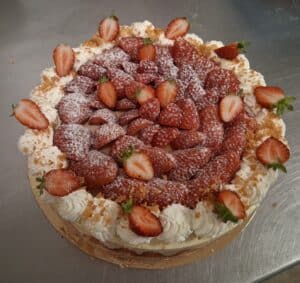 Gâteau rond généreusement garni de fraises fraîches coupées, saupoudré de sucre glace et entouré de crème chantilly et de miettes de biscuits.