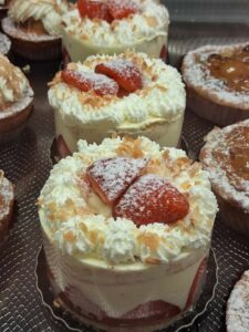 Trois petits gâteaux individuels à la crème et aux fraises coupées, décorés de copeaux de noix de coco ou d'amandes effilées, présentés sur un étal de pâtisserie.