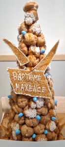 Pièce montée de baptême en choux caramélisés et nougatine, décorée de perles bleues et d'un panneau en nougatine portant l'inscription "BAPTÊME MAXENCE" en glaçage blanc.