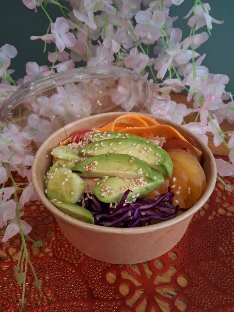 Poke Bowl Frais : Avocat, Chou Rouge et Légumes Croquants Poke bowl frais avec avocat, concombre, chou rouge et sésame dans un bol kraft devant des fleurs de cerisier.